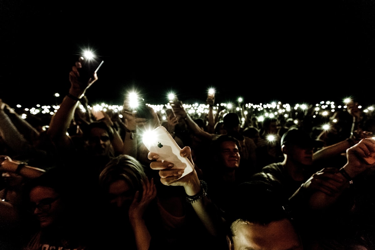 A sea of phone screens glowing in darkness — technology demanding our attention